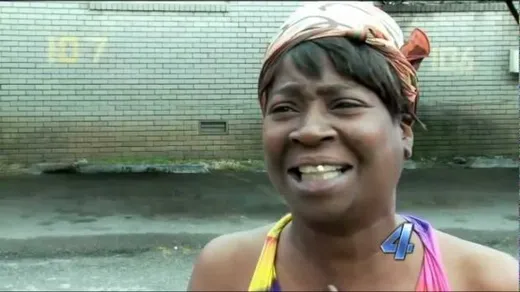 A woman in a head wrap talking animatedly to a news camera outdoors