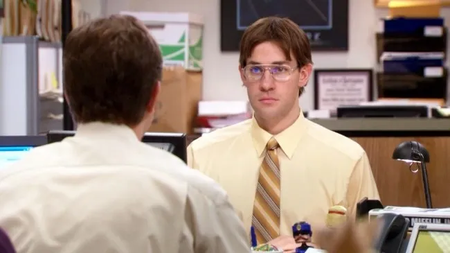 Jim Halpert in glasses, a mustard shirt and a side parting, imitating Dwight at his desk