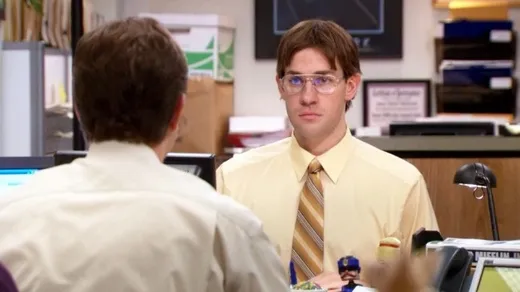 Jim Halpert in glasses, a mustard shirt and a side parting, imitating Dwight at his desk