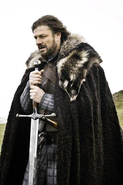 Ned Stark in fur-trimmed armour holding a large sword, looking grim