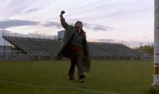 A young man in a jacket on a football field, raising his fist in the air