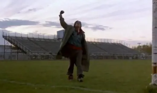 A young man in a jacket on a football field, raising his fist in the air