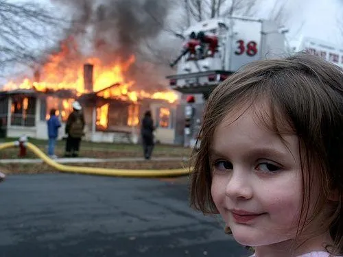 A young girl smiling knowingly at the camera, with a house on fire and firefighters behind her
