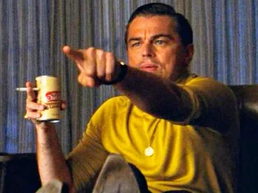 Rick Dalton in a yellow shirt, leaning forward in an armchair, holding a beer and pointing