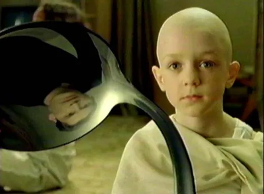 A bald boy in robes holding a bent spoon, reflected in the bowl