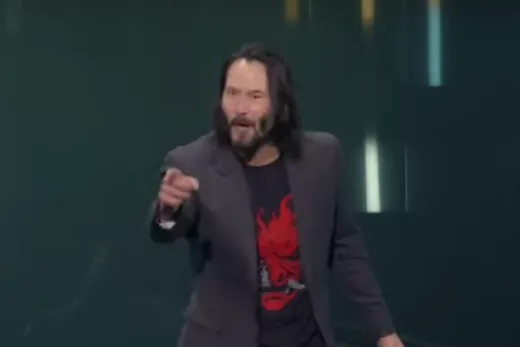 Keanu Reeves in a black suit on stage, pointing at the audience and smiling